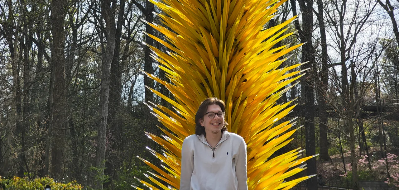 A person with shoulder-length brown hair and glasses is looking towards the camera and smiling. They are wearing a cream quarter zip turtleneck (unzipped). They are sitting at the bottom of the frame. Behind them is a large sculpture, a tall pillar with yellow glass spikes similar to a pine cone. Behind the pillar are several trees with their spring leaves just beginning to grow.